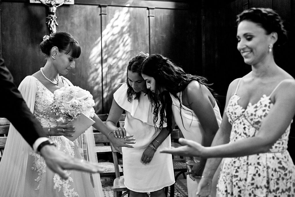 Cérémonie à l'église - plusieurs niveaux de lecture du plan : premier plan la main du marié avec alliance, et arrière plan la mariée avec sa main qui montre son alliance à ses copines - photographe mariage style reportage